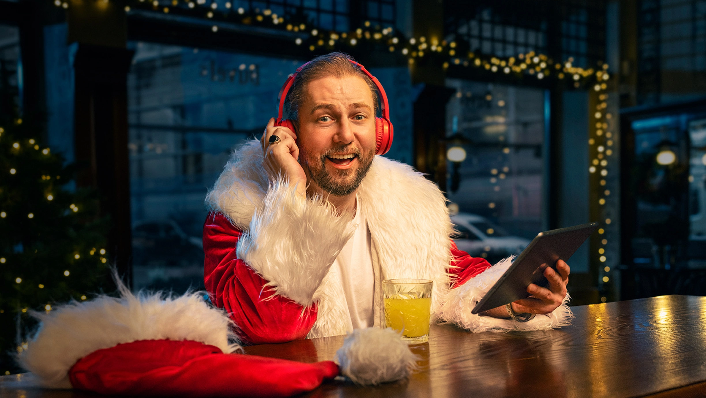 Vīrietis Ziemassvētku vecīša tērpā ar sarkanām austiņām pie letes, tur planšeti; fonā svētku gaismiņas.