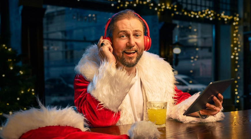 Vīrietis Ziemassvētku vecīša tērpā ar sarkanām austiņām pie letes, tur planšeti; fonā svētku gaismiņas.