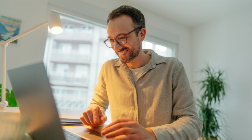 Smaidošs vīrietis strādā pie datora, blakus galda lampa un telpaugs