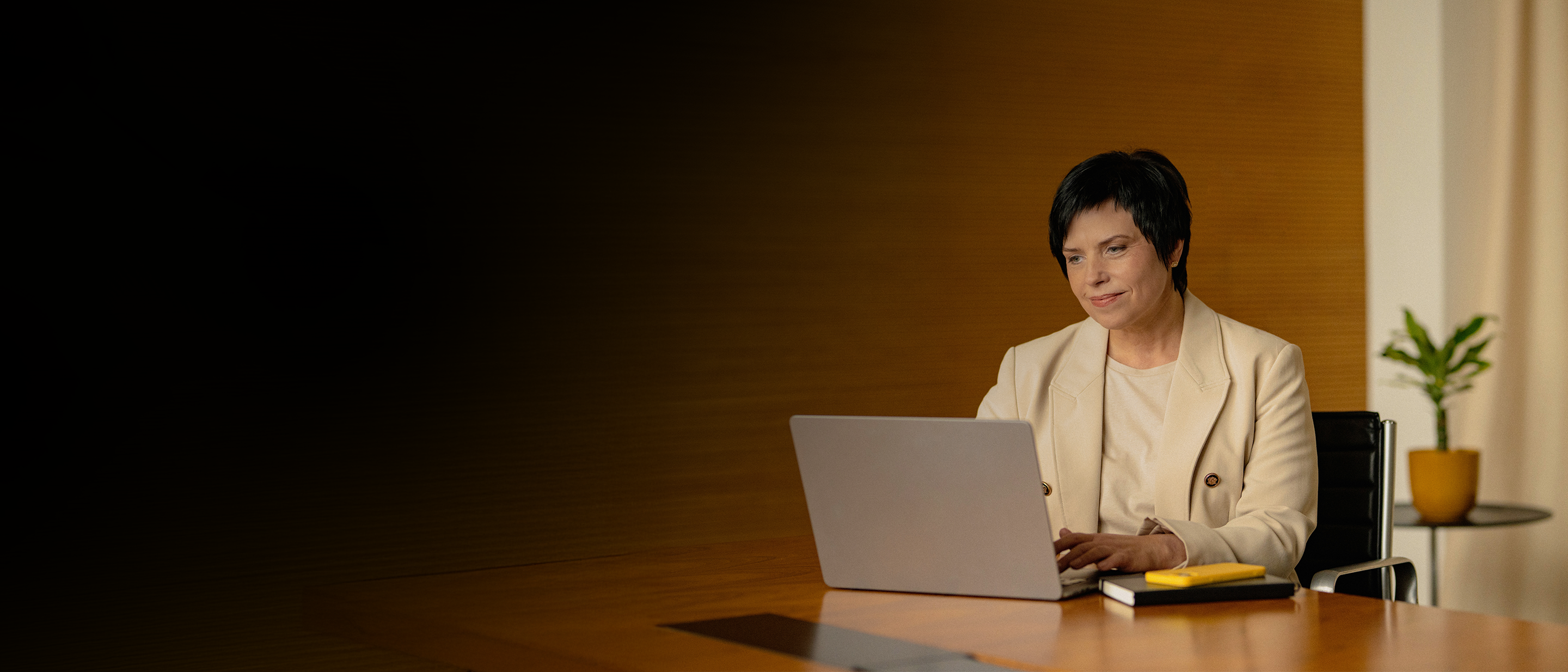 Sieviete gaišā jakā pie koka galda strādā ar klēpjdatoru; fonā telpaugs.