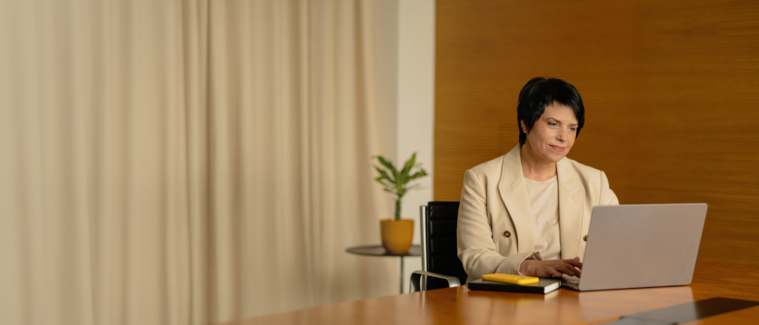 Sieviete ar tumšiem matiem bēšā žaketē sēž pie galda un skatās portatīvajā datorā