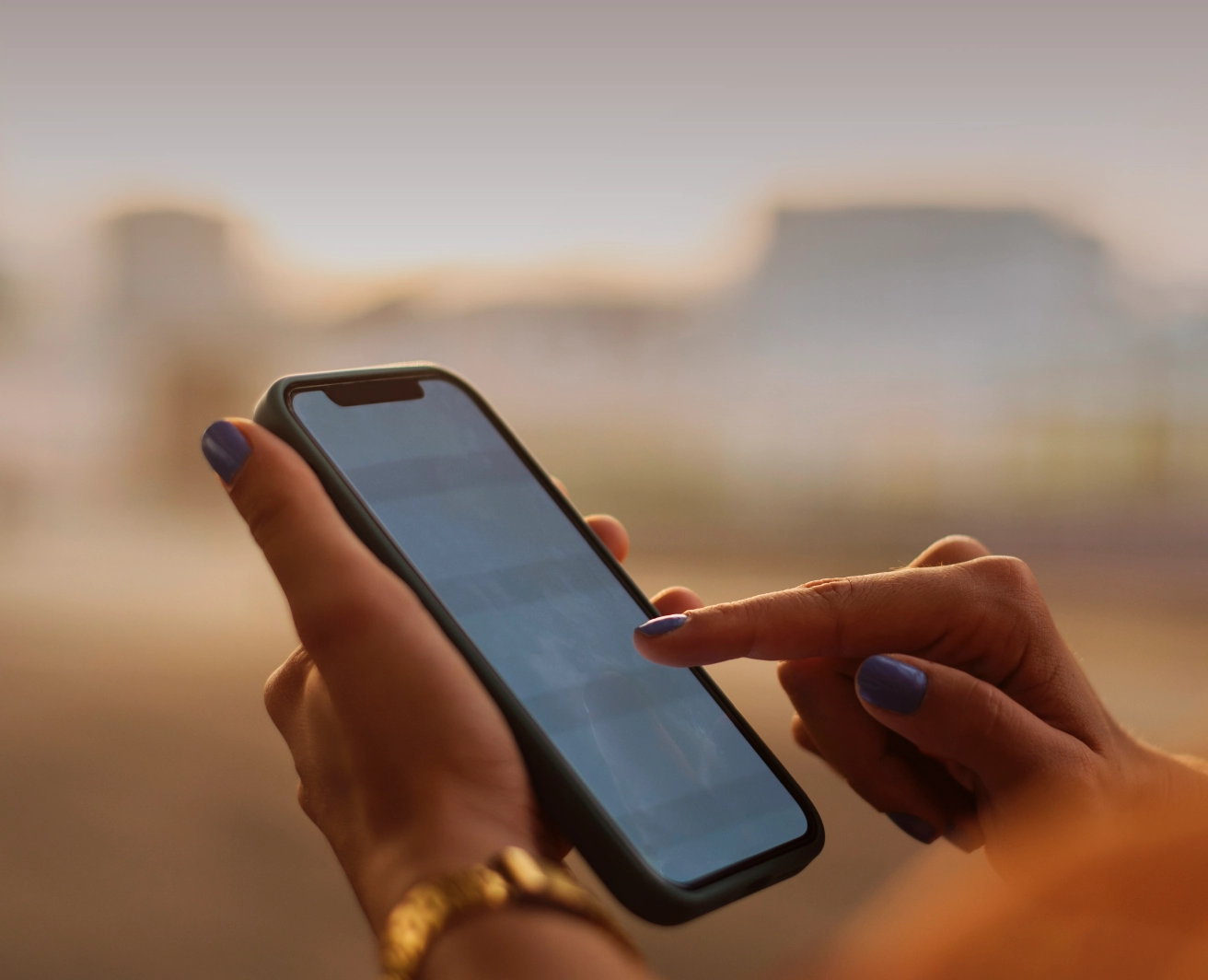 Tuvplāns ar cilvēka rokām, kas tur melnu mobilo telefonu un pieskars tā ekrānam, fonā izplūdis silts apgaismojums, cilvēkam ar roku zelta krāsas pulkstenis.