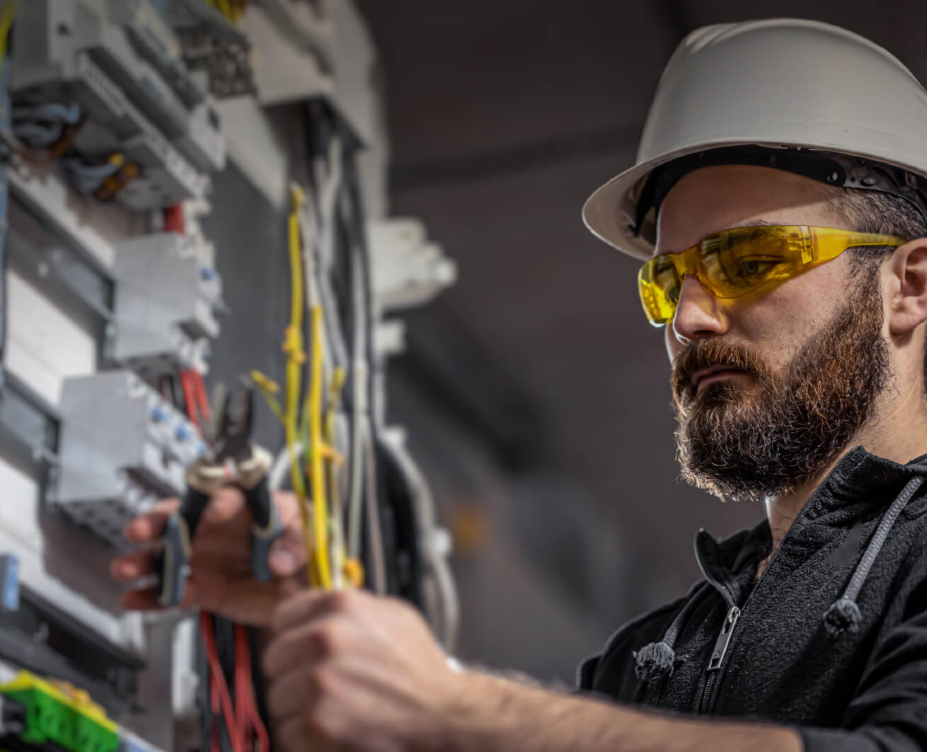 Elektriķis ar ķiveri un dzeltenām aizsargbrillēm strādā pie sadales skapja. Rokā knaibles, tiek kārtoti dzelteni un sarkani vadi; drošības aprīkojums, koncentrēts skatiens.
