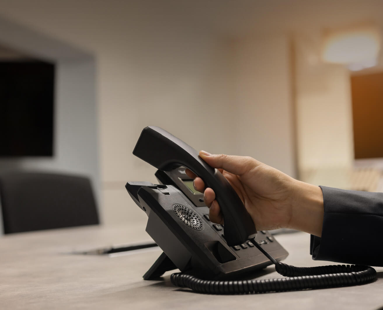 Roka paceļ galda telefona klausuli biroja telpā. Uz galda telefons ar spirālveida vadu; fonā sapulču telpas krēsli un izpludināts interjers.