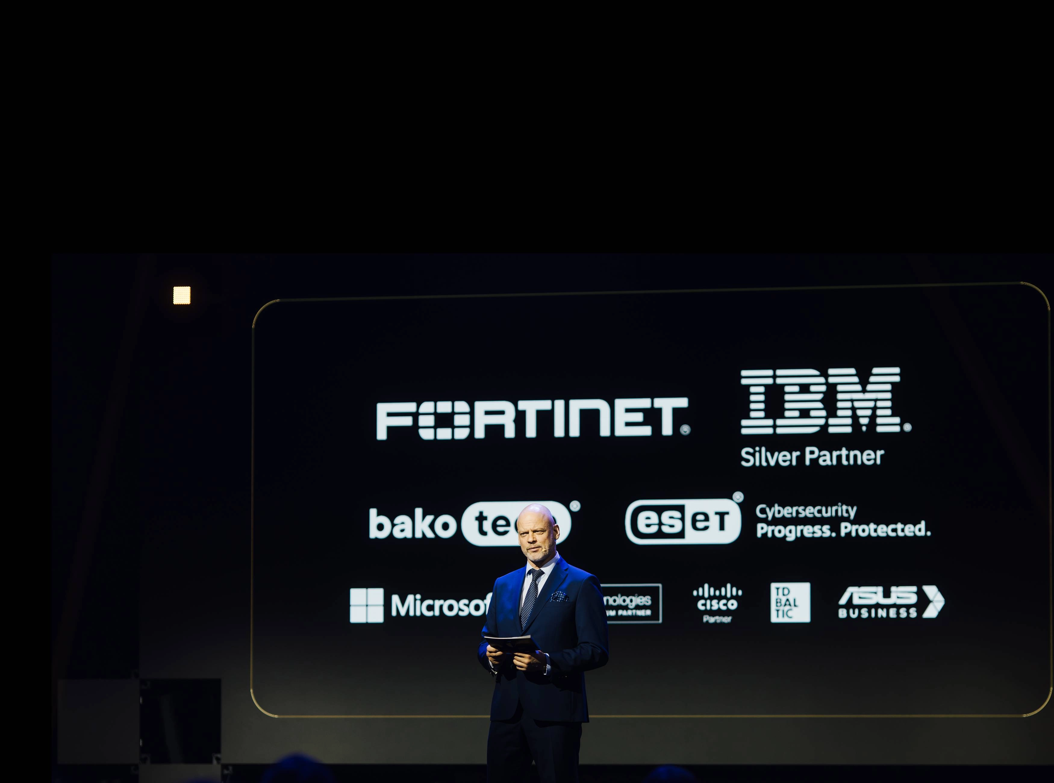 Konferences skatuve ar vadītāju, kurš tur planšeti, fonā lielo tehnoloģiju un kiberdrošības uzņēmumu logo, tostarp Fortinet, IBM, ESET, Microsoft, Cisco un ASUS Business