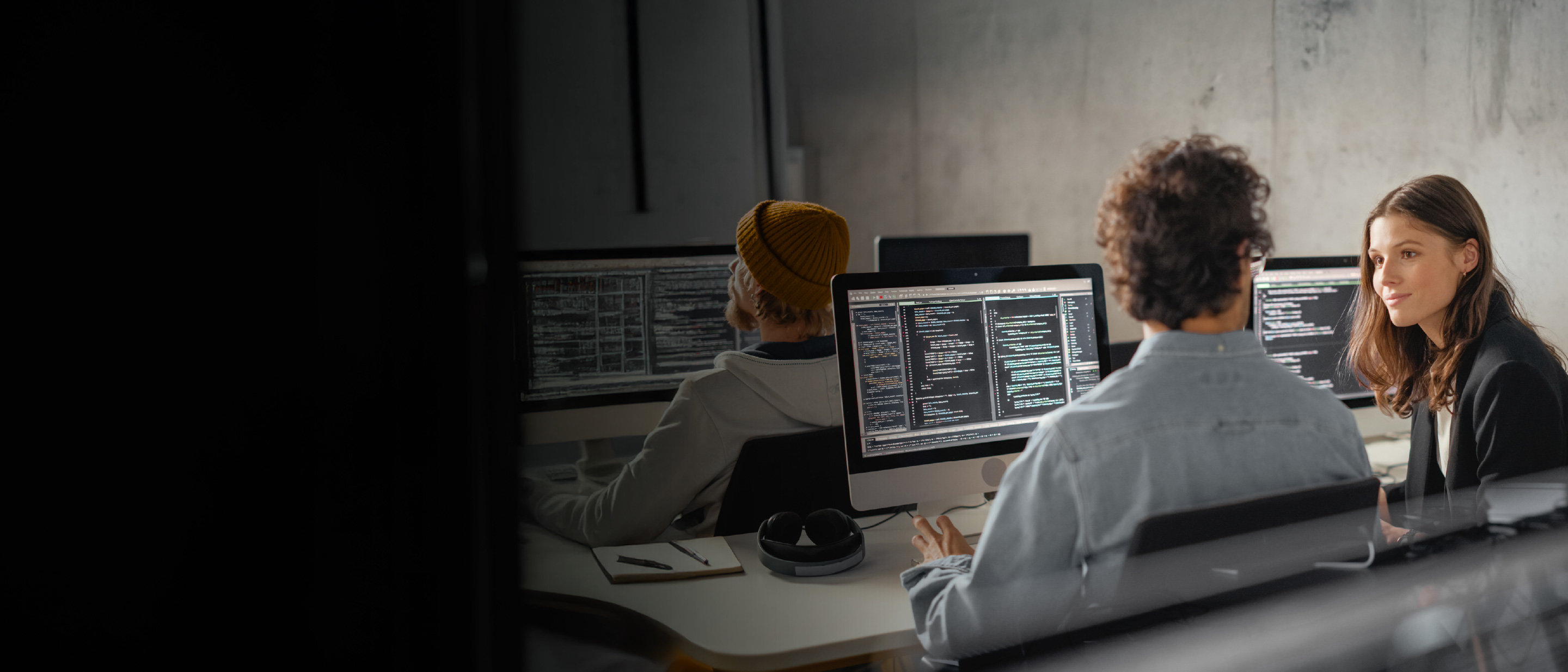 Trīs cilvēki birojā strādā pie monitoriem ar programmas kodu. Priekšplānā divi kolēģi pārrunā risinājumu, blakus austiņas un piezīmju bloks; fonā vēl viens programmētājs ar dzeltenu cepuri.