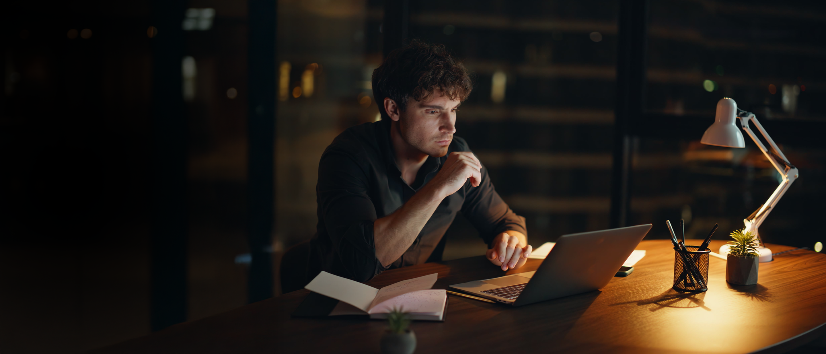 Vīrietis vēlu vakarā strādā pie klēpjdatora birojā; lampa, klade un mazs augs uz galda.