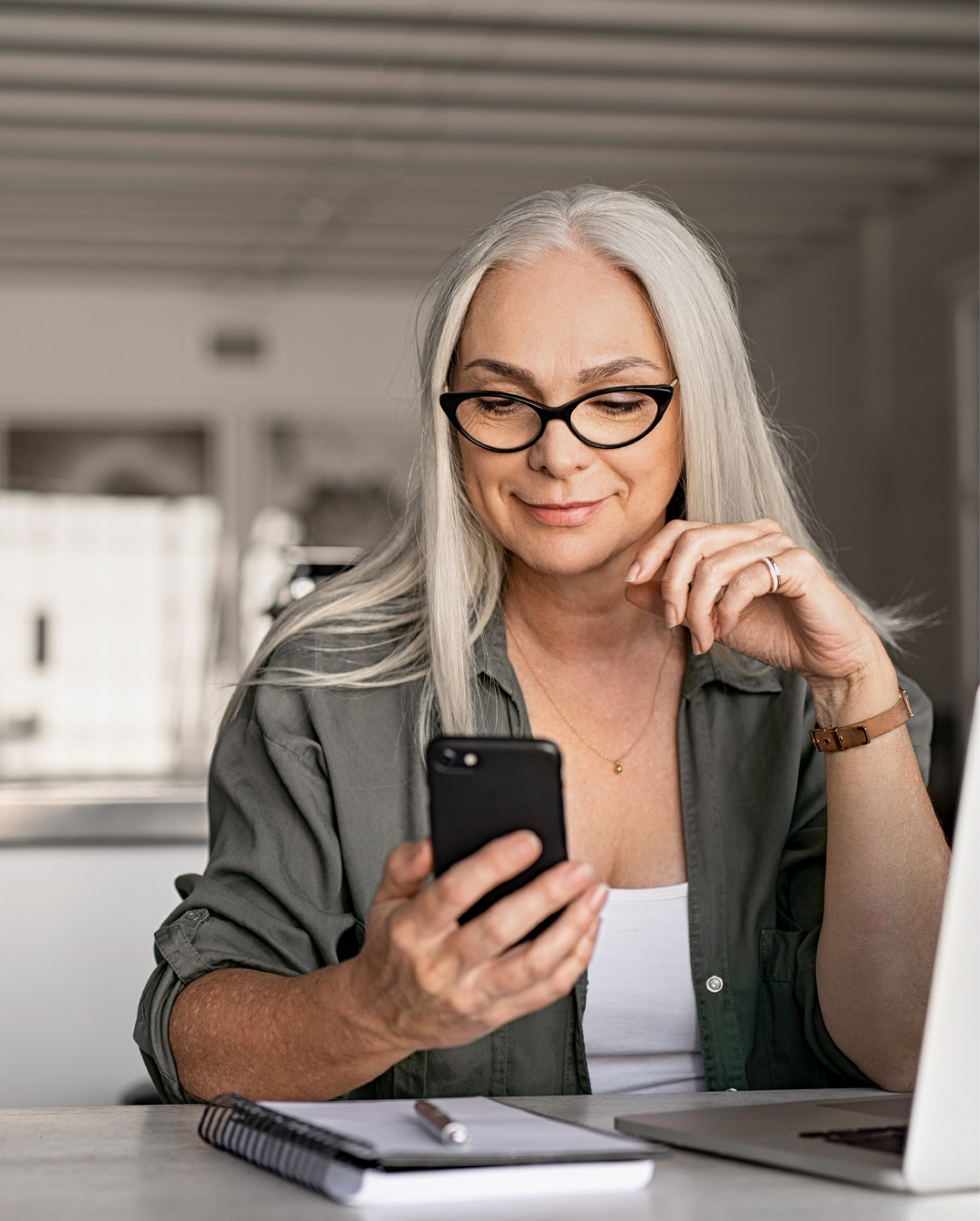 Smaidīga persona ar sirmiem matiem lieto viedtālruni pie klēpjdatora.