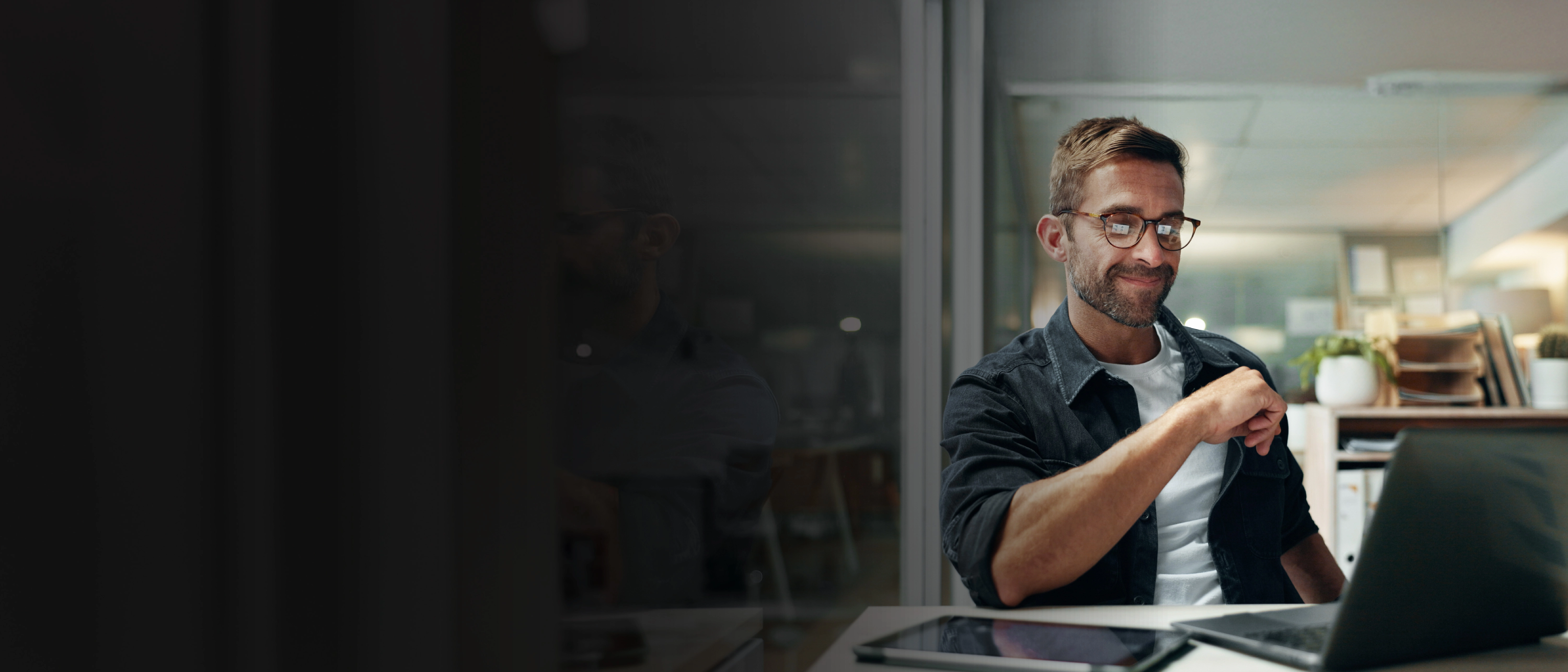 Persona ar brillēm vakara birojā skatās klēpjdatorā; priekšplānā planšete un dokumenti.