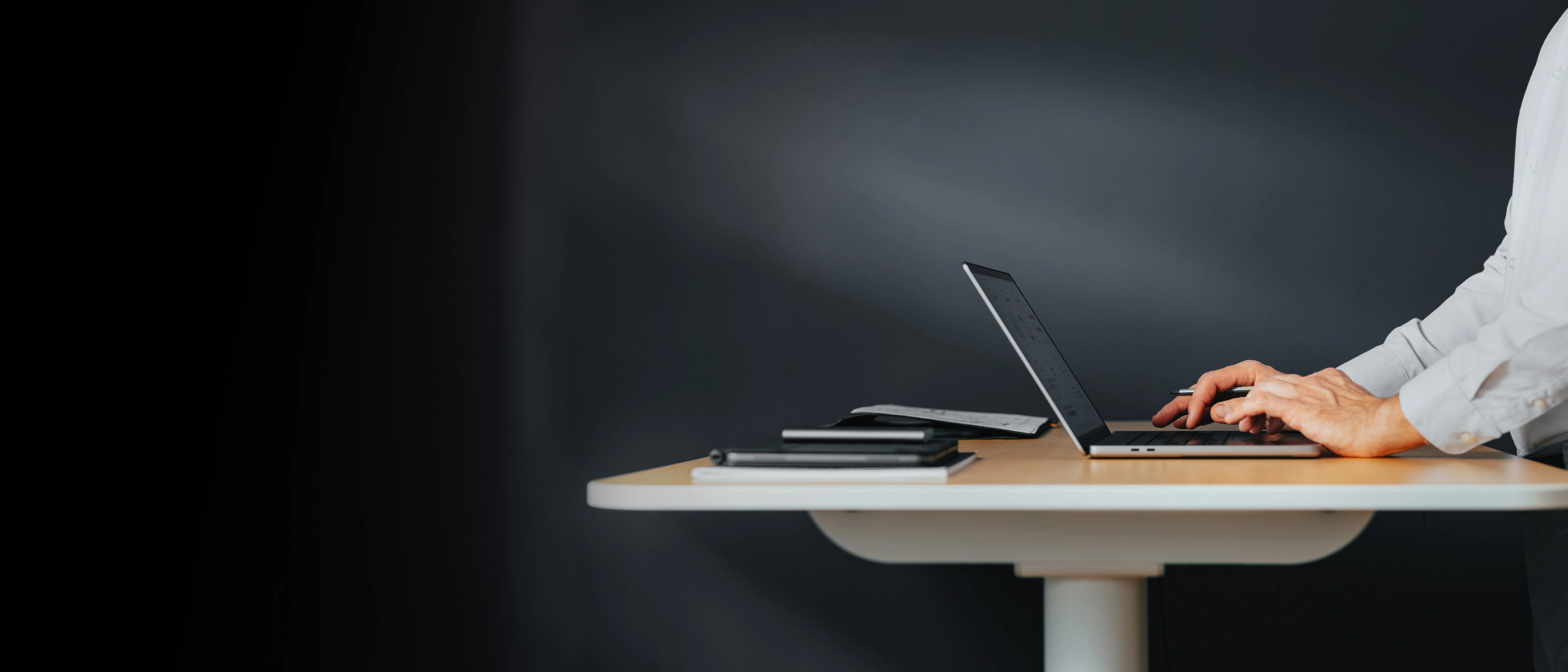 Rokas raksta uz klēpjdatora pie stāvgalda, tumšs fons.