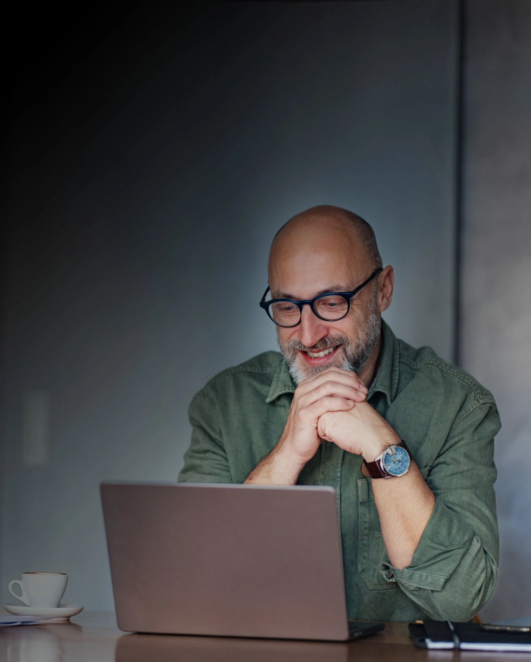 Vīrietis ar brillēm smaida, skatoties portatīvajā datorā.