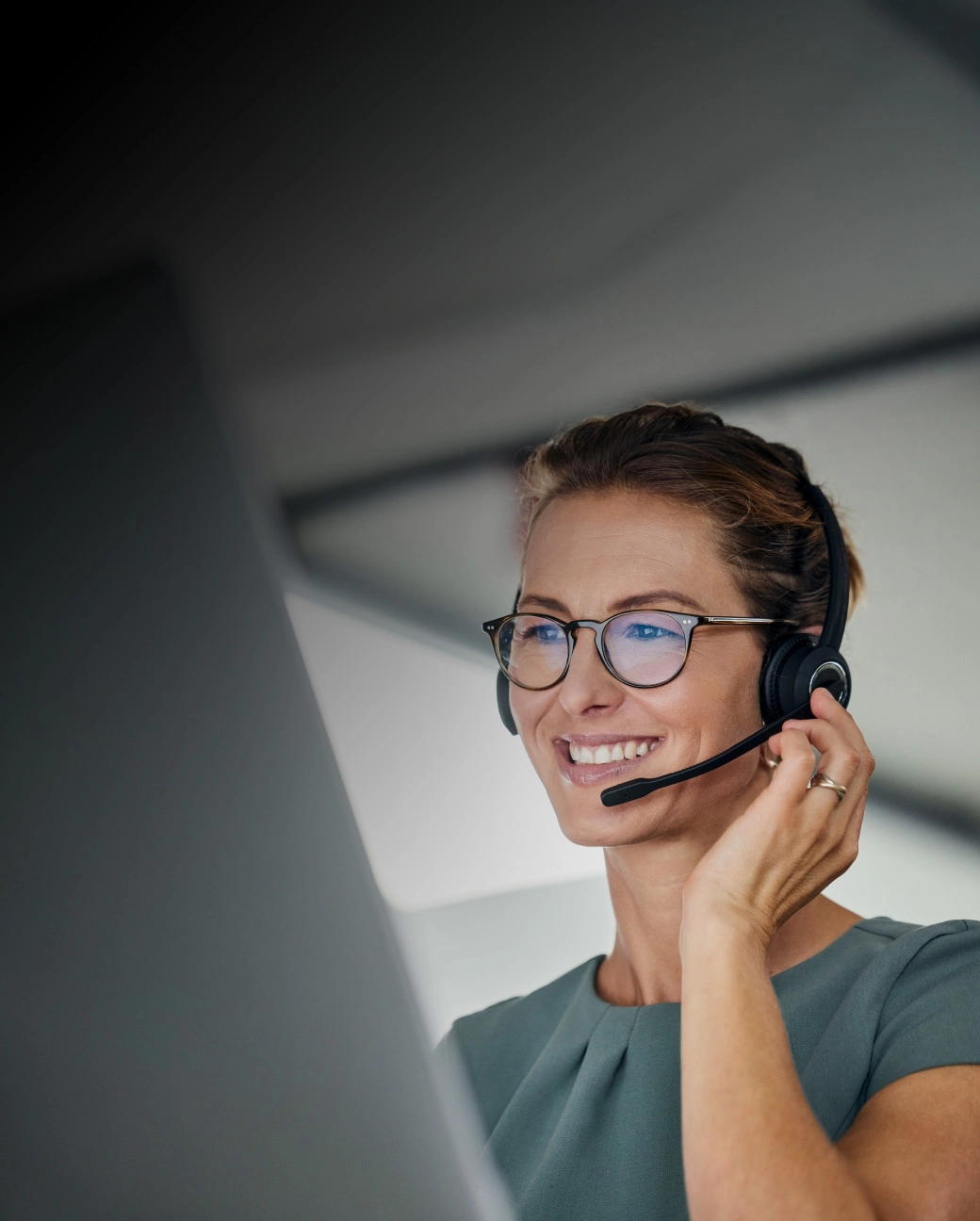 Sieviete ar austiņām un mikrofonu smaidot skatās datorā.