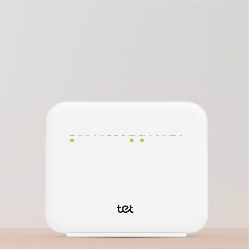 Balts Wi-Fi maršrutētājs ar zaļām indikatora gaismām priekšpusē, novietots uz gaiša koka virsmas. Uz ierīces zīmols Tet.
