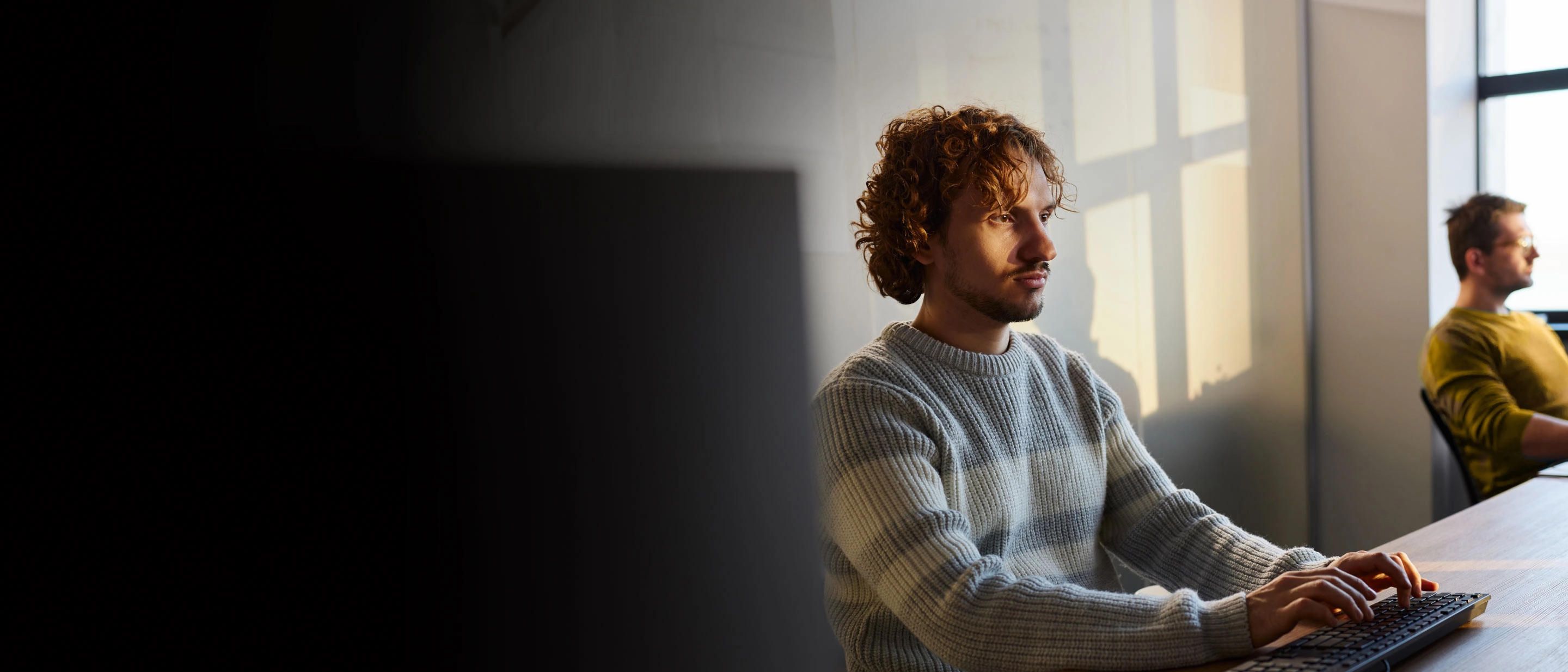 Jauns vīrietis ar cirtainiem matiem strādā pie datora gaišā telpā, fonā vēl viens cilvēks. Saules gaisma krīt pa logu.
