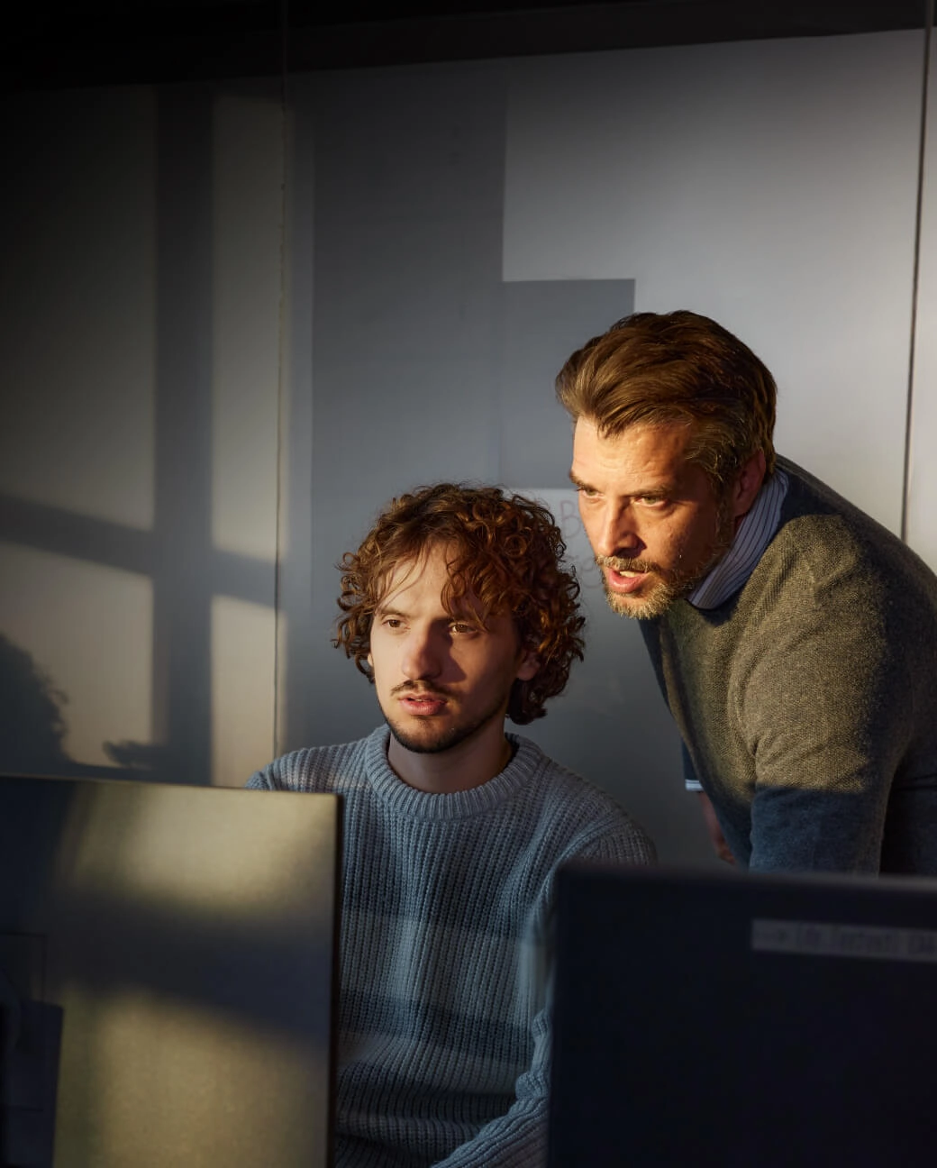 Divi vīrieši birojā koncentrēti skatās datora ekrānā, viens sēž, otrs stāv blakus un palīdz risināt problēmu.