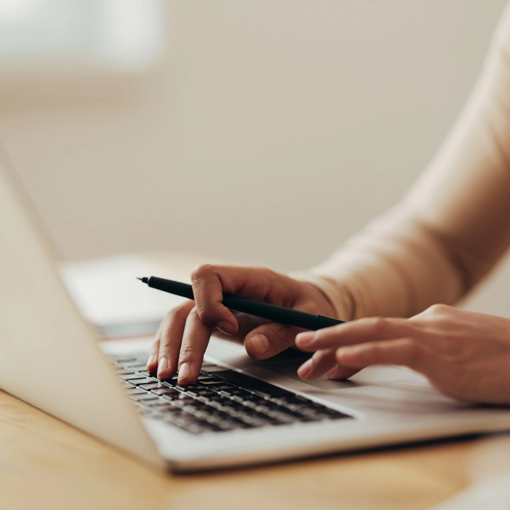 Roka tur pildspalvu un otrā roka raksta uz klēpjdatora tastatūras, gaiša un mājīga vide.