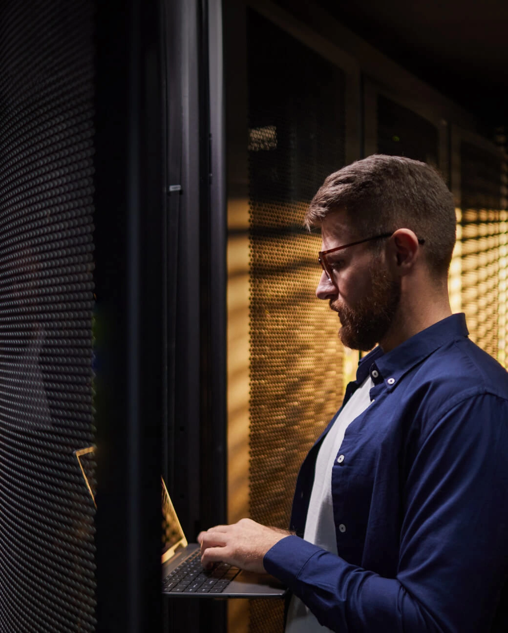 Vīrietis ar brillēm un bārdu strādā datu centrā pie portatīvā datora, fonā serveru statīvi ar apgaismojumu.