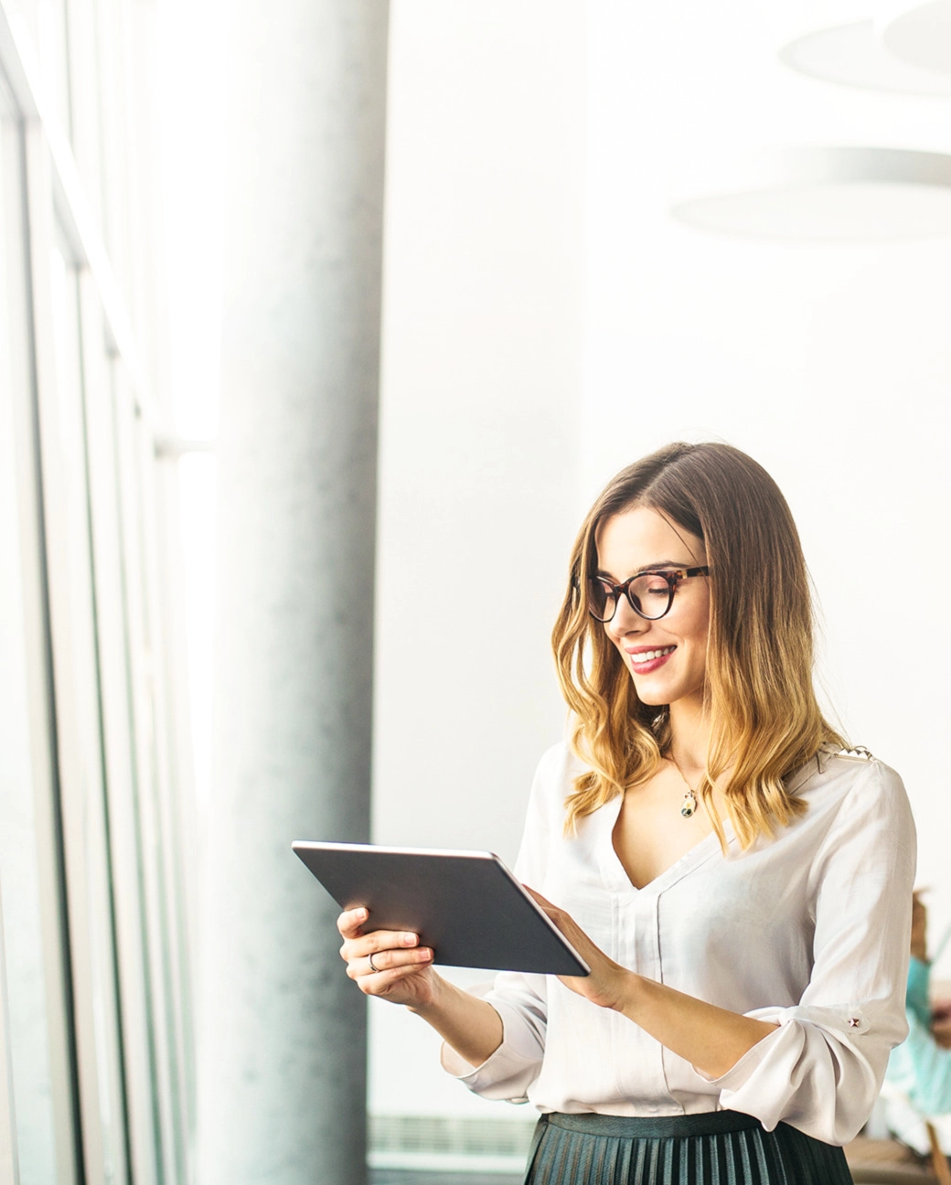 Sieviete ar brillēm, gaišā blūzē smaida un skatās planšetdatorā, atrodas gaišā, modernā telpā pie loga.