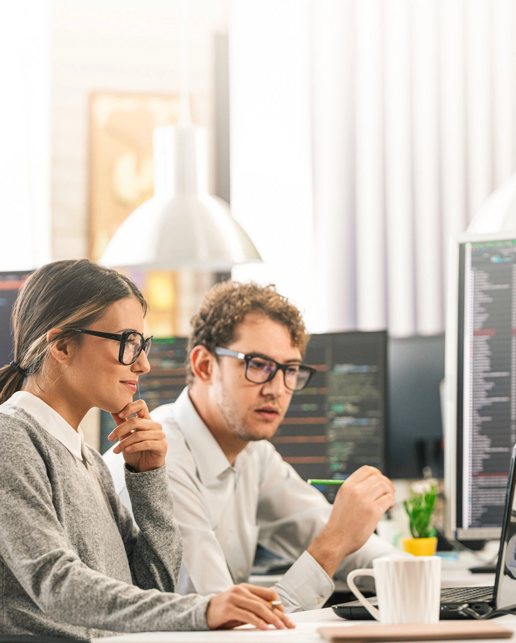 Divi kolēģi strādā birojā pie datora monitoriem ar kodu, koncentrējušies uz darbu, uz galda balta kafijas krūze.