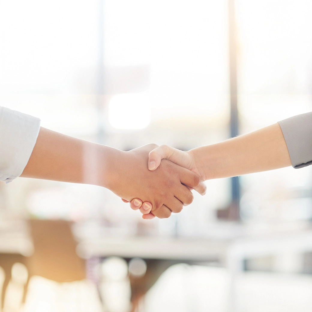 Divi cilvēki paspiež viens otram roku gaišā, mūsdienīgā birojā, aizmugurē neasa mēbeļu kontūra un silta gaisma.