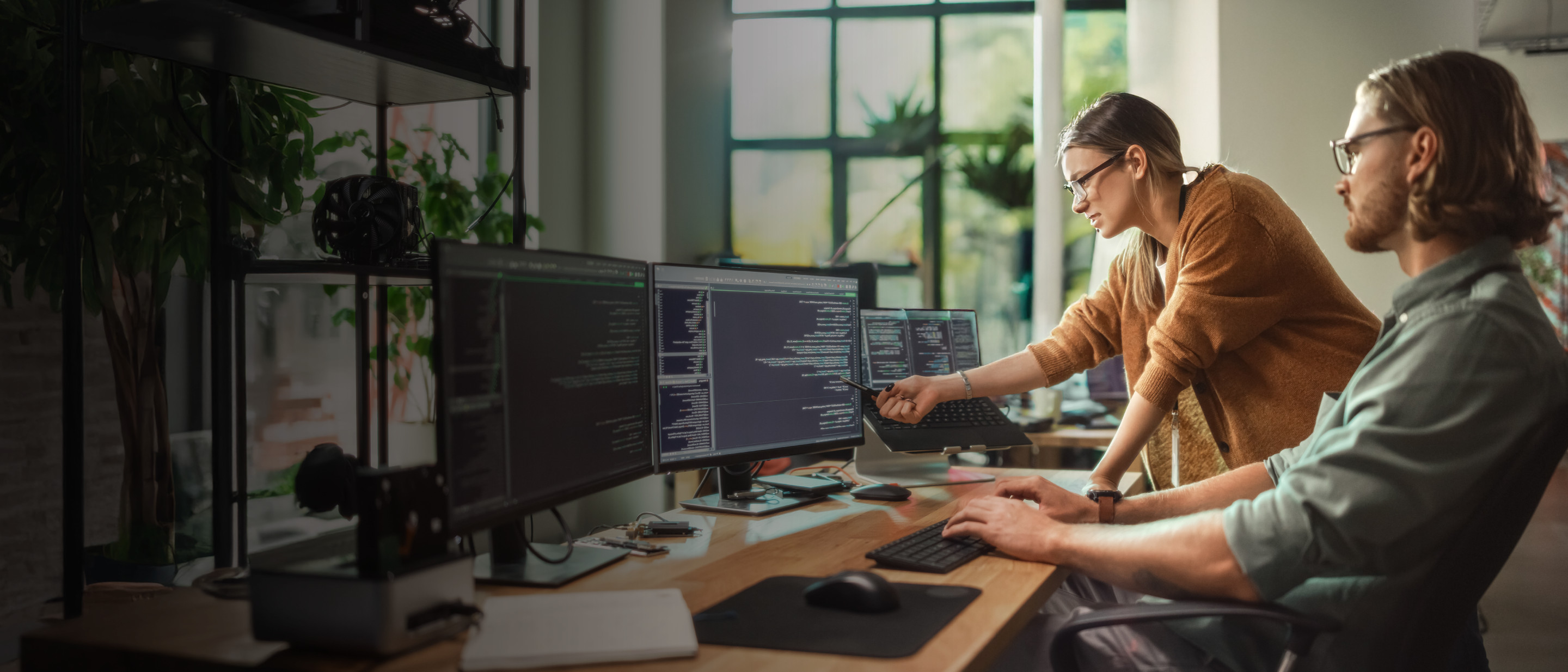 Divi cilvēki birojā strādā pie vairākiem monitora ekrāniem ar programmēšanas kodu, sieviete rāda uz ekrānu, vīrietis raksta uz tastatūras.