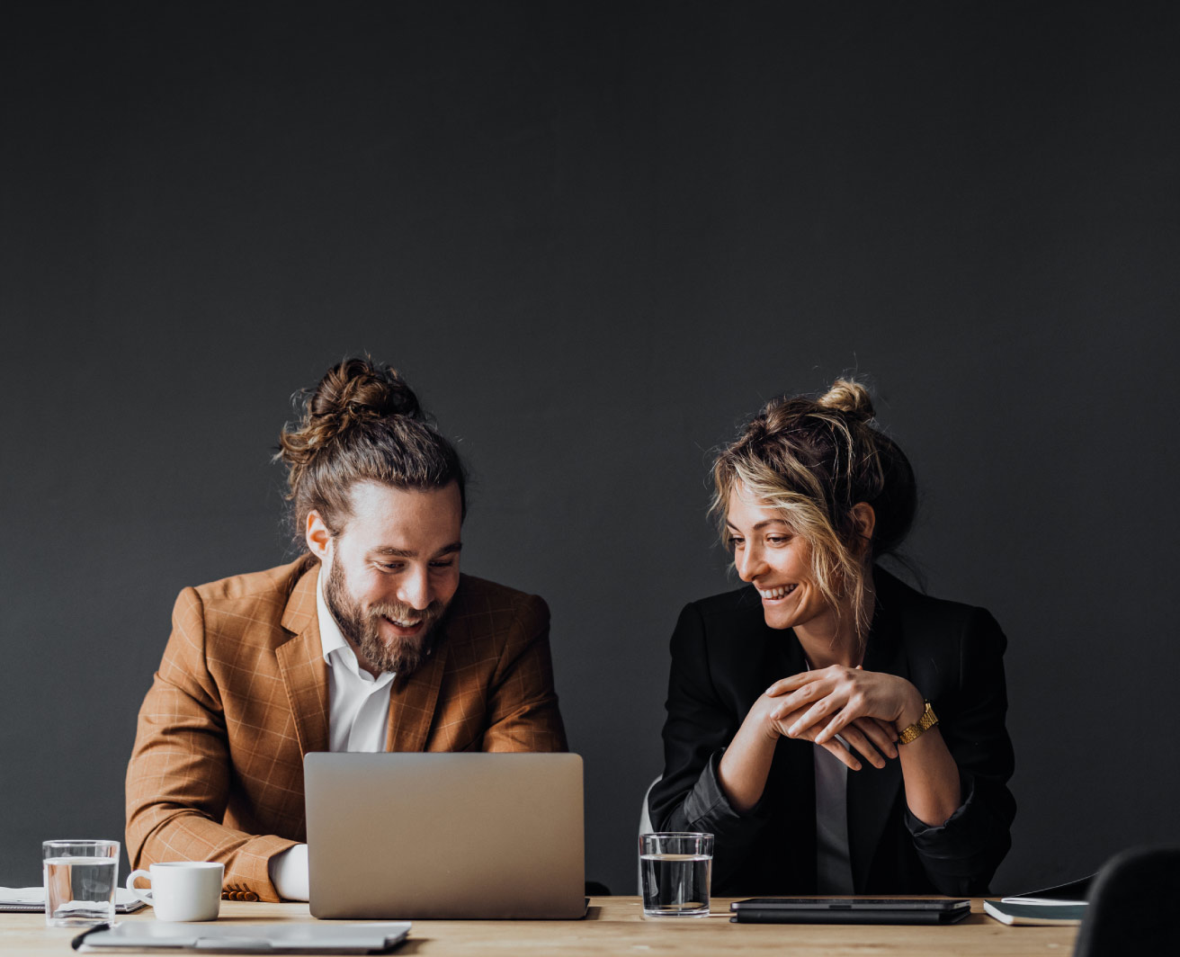 Smaidīgi kolēģi kopā strādā pie klēpjdatora pie galda, uz tumša minimālistiska fona.