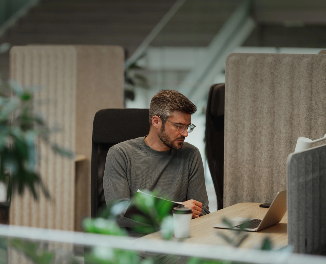 Vīrietis sēž pie biroja galda ar atvērtu portatīvo datoru, vīrietim rokās ir dokumenti, bet uz galda kafijas krūze; fonā mūsdienīga biroja interjers ar zaļiem augiem.