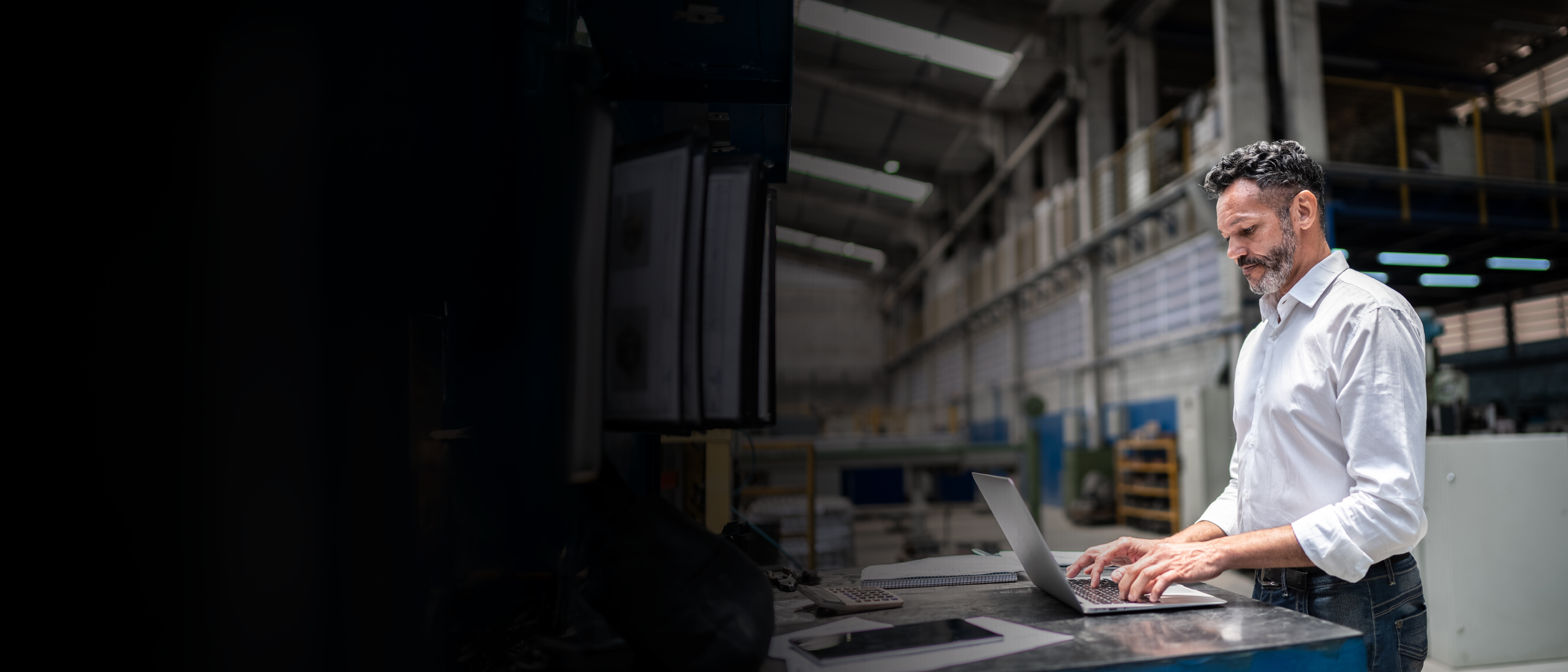 Vīrietis industriālā cehā raksta klēpjdatorā pie darba virsmas ar dokumentiem; fonā ražošanas telpa.