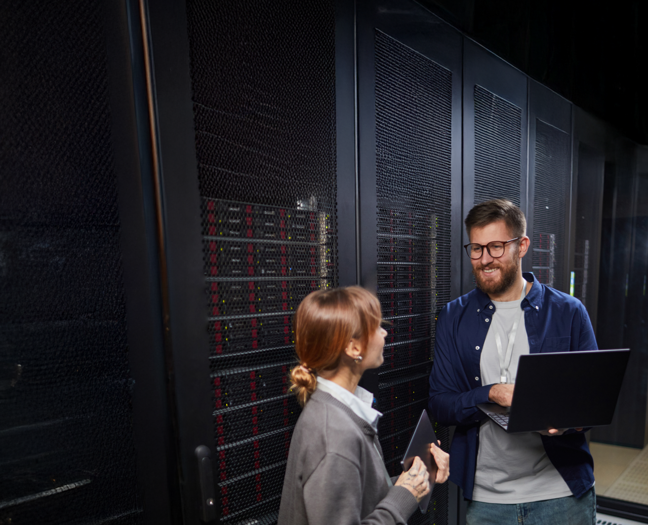 Divi IT speciālisti serveru telpā: vīrietis ar klēpjdatoru un sieviete ar planšeti pie serveru skapjiem.