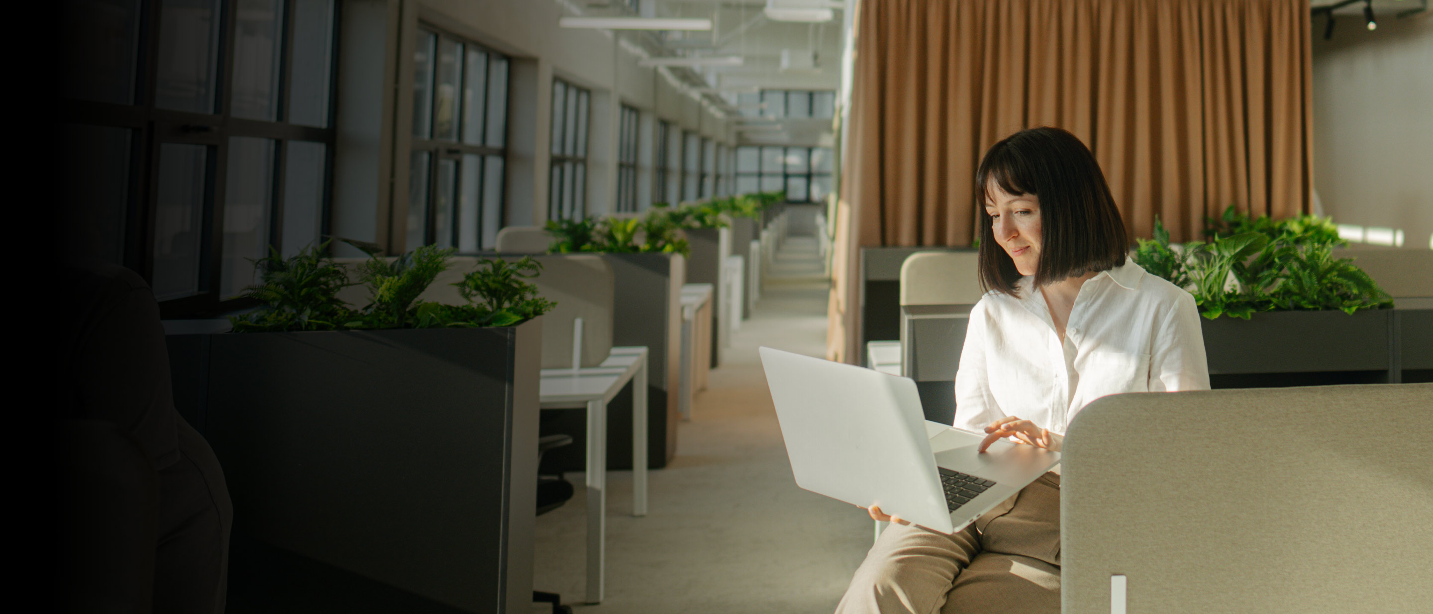 Saulaina biroja telpa ar rindā izkārtotām darbvietām un augiem; priekšplānā persona ar klēpjdatoru.