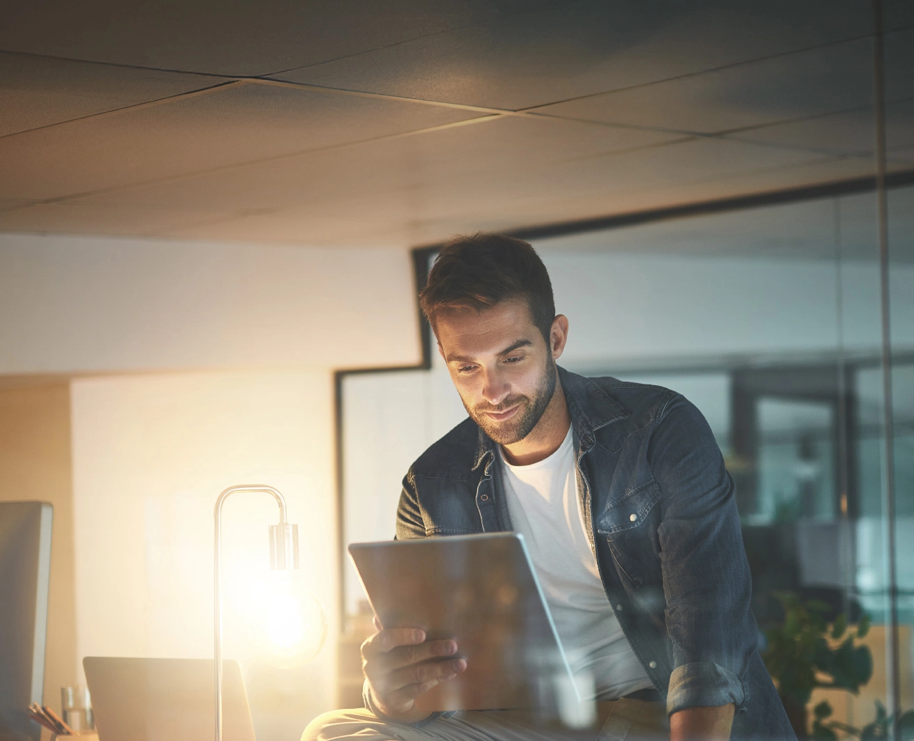 Vīrietis, tumši zilā džinsa kreklā zem kura ir balts t-krekls, sēž uz galda biroja vidē un tur rokās planšetdatoru, blakus galda lampa ar spožu, siltu apgaismojumu.