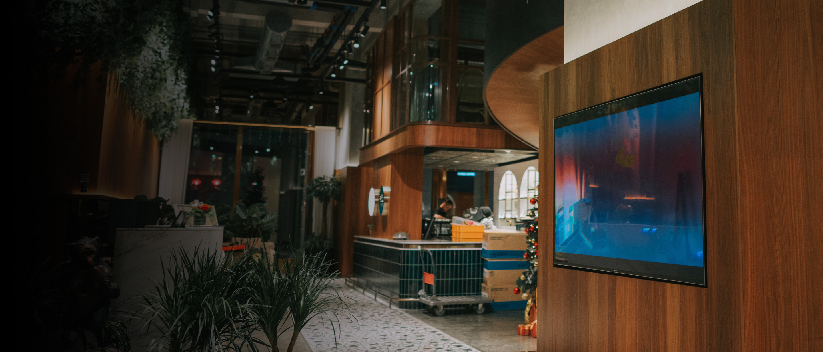  Plakanais TV koka paneļa sienā, fonā restorāna/foajē zona ar augiem un dekorācijām.