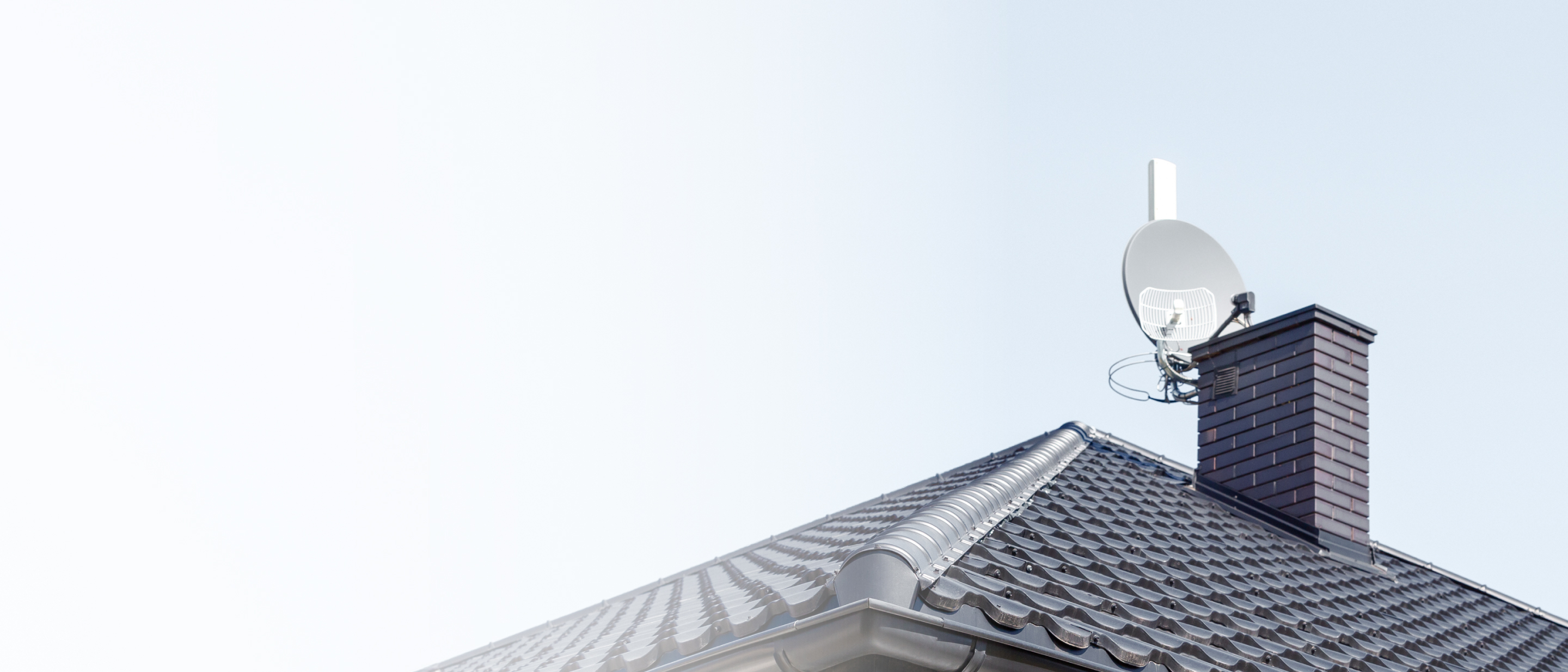 Pelēks dakstiņu jumts ar dūmeni un satelīttelevīzijas antenu pret skaidrām debesīm.