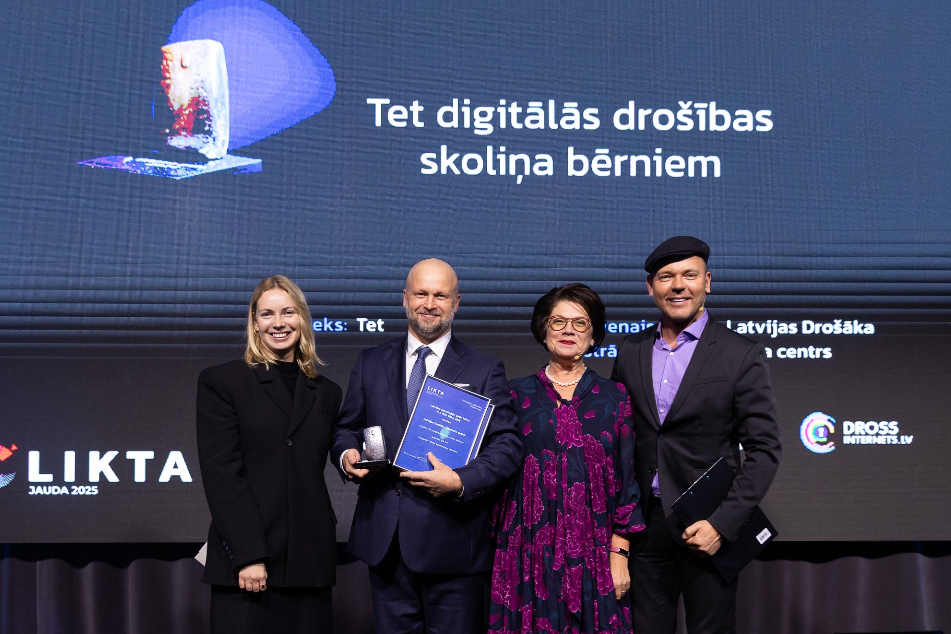 Četri cilvēki uz skatuves pozē ar apbalvojumu; fonā ekrāns ar tekstu “Tet digitālās drošības skoliņa bērniem” un LIKTA, Dross Internets logotipiem.