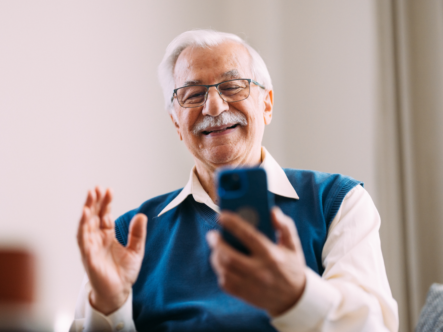 Priecīgs kungs ar brillēm skatās viedtālrunī un sveicina kameru.