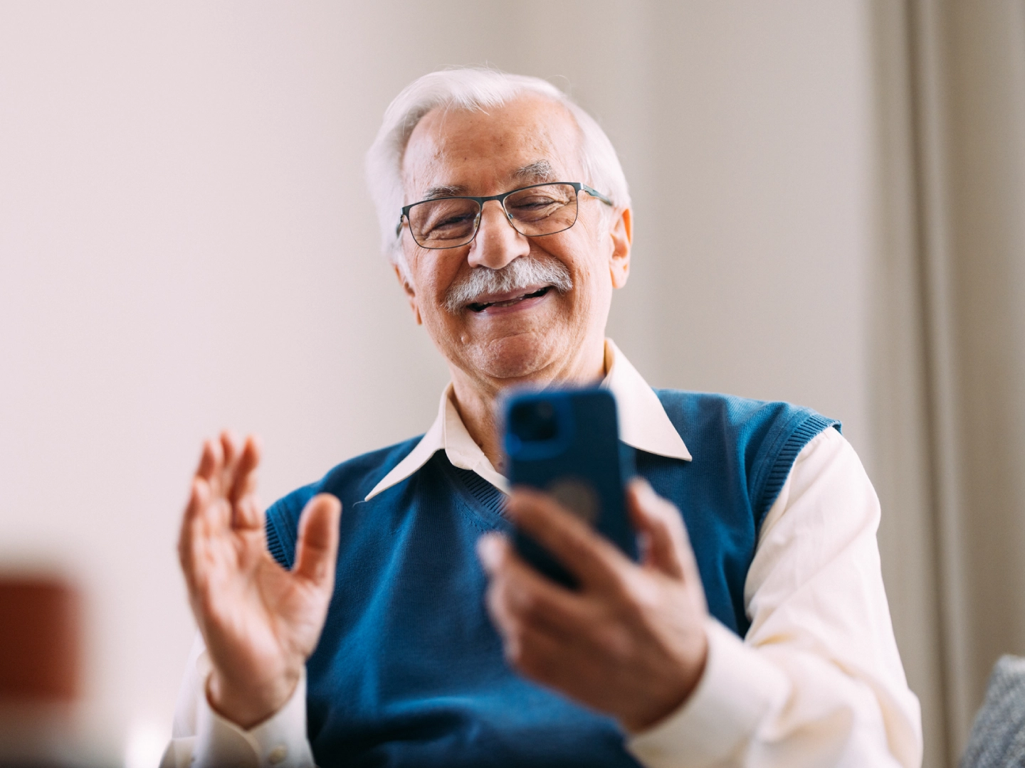 Priecīgs kungs ar brillēm skatās viedtālrunī un sveicina kameru.