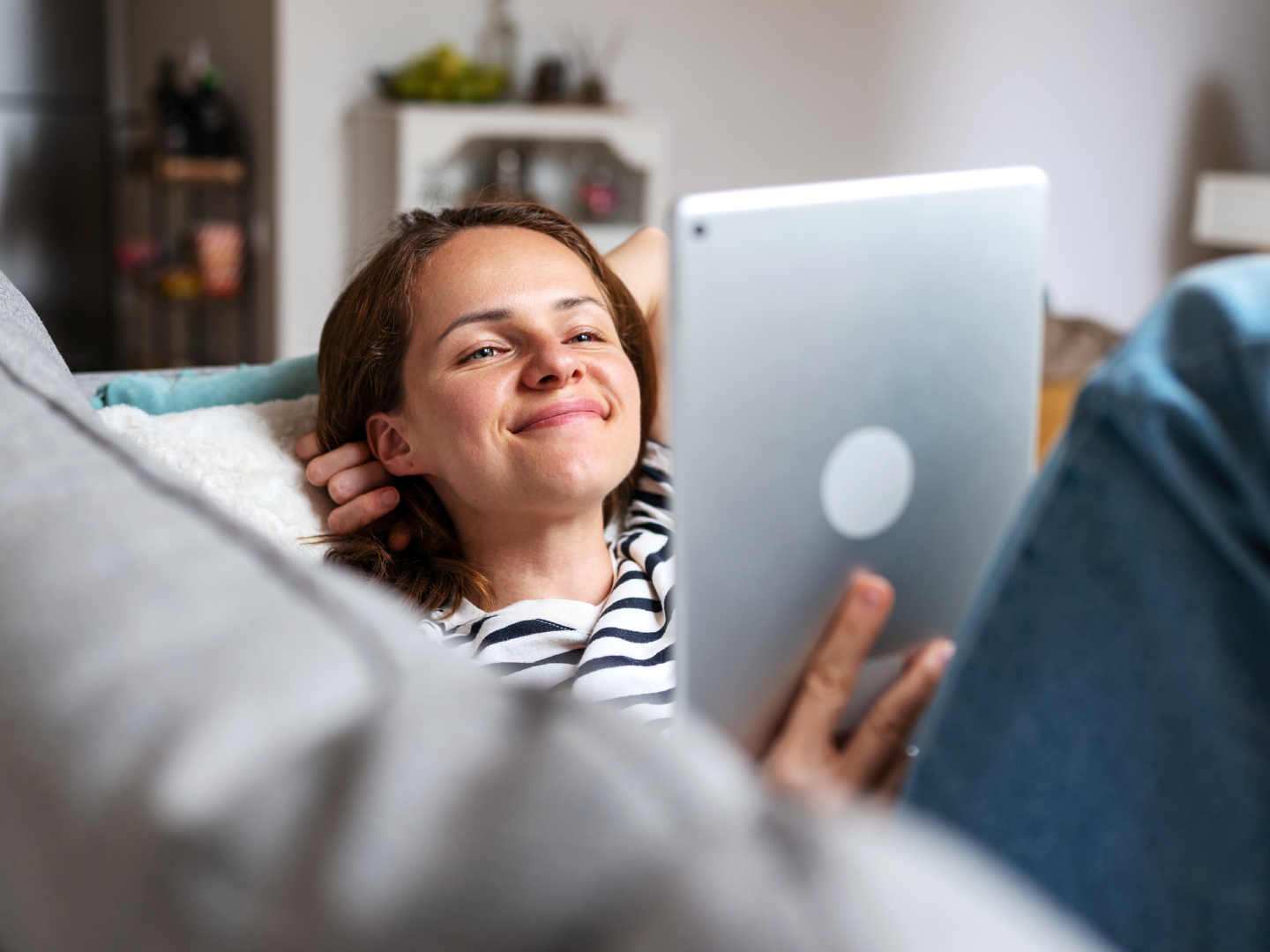 Sieviete atslābusi dīvānā smaidot skatās planšetē mājās.